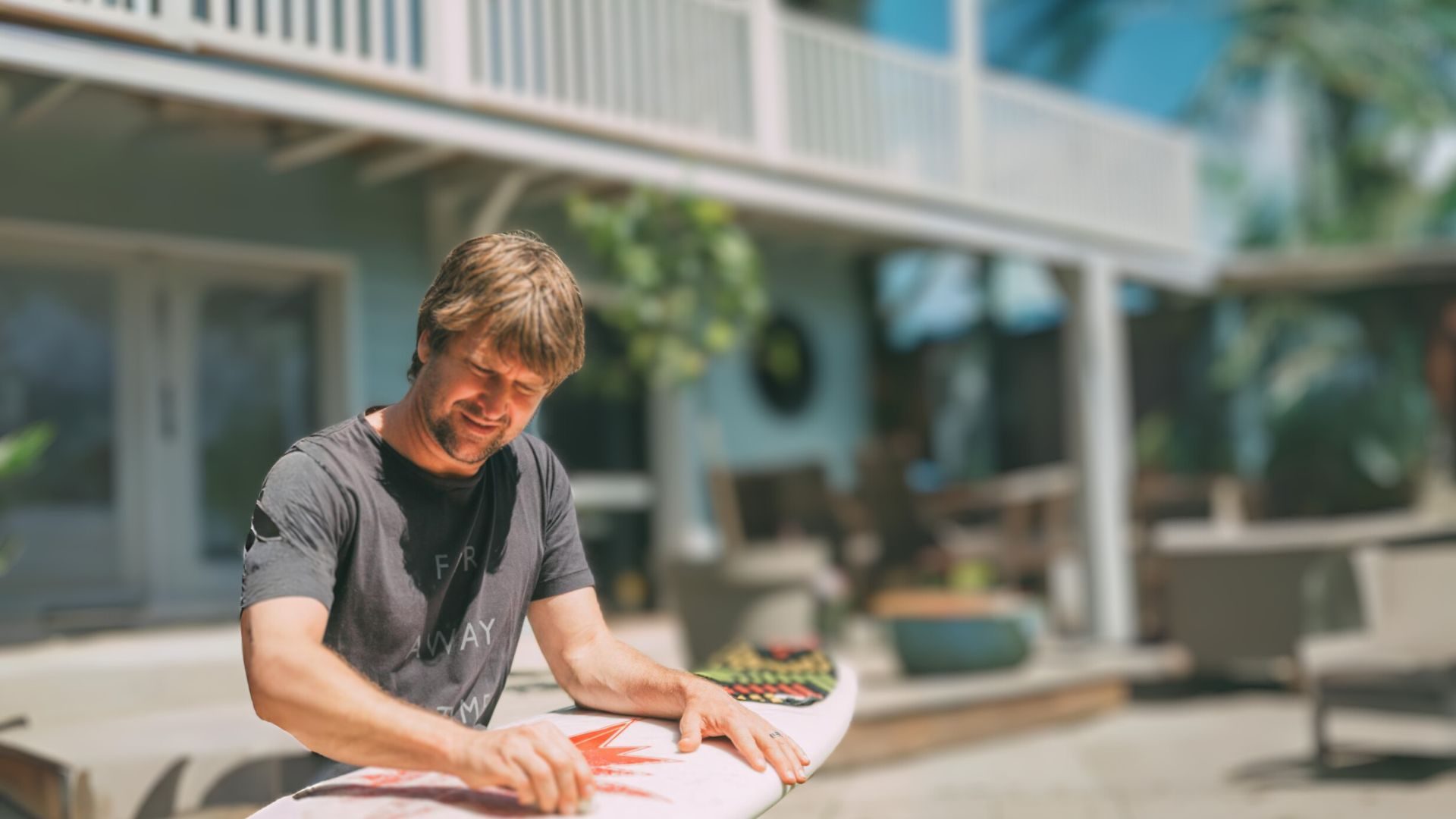 Tom Frager en train de waxer sa planche de surf, en arrière plan sa maison bleu