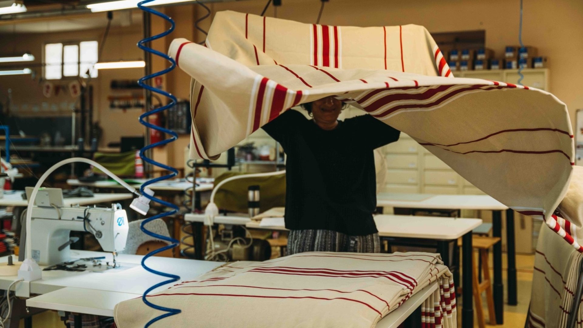 Artisan textile travaillant dans un atelier, soulevant un grand tissu à rayures rouges et blanches avec une machine à coudre industrielle en arrière-plan